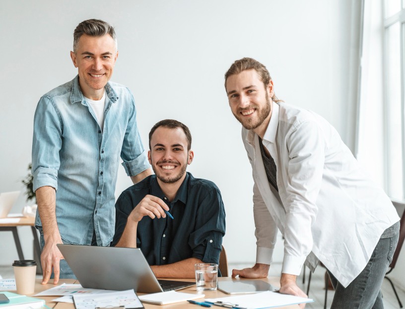 Drei Kollegen lachen gemeinsam im Büro am Laptop – freundliche Arbeitsatmosphäre bei Rechtsschutzengel.