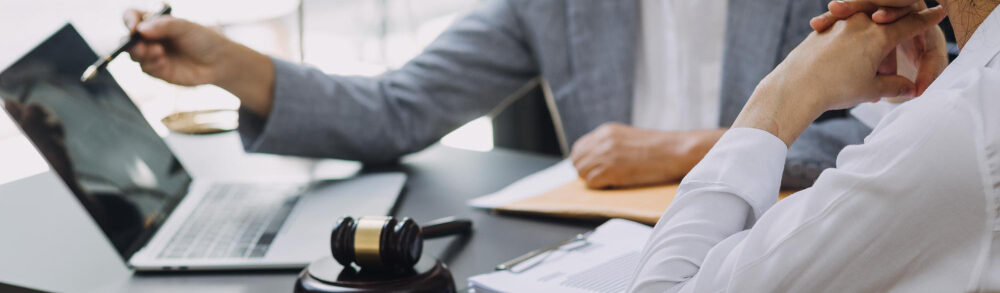 Zwei Personen im Gespräch am Schreibtisch mit Laptop, Richterhammer und Vertragsunterlagen – Zusammenarbeit mit Kanzleien und Beratern.